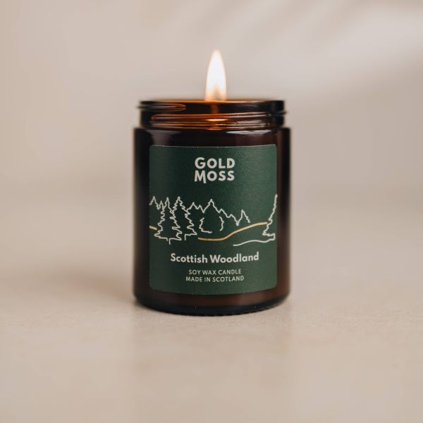 A lit amber glass jar candle labeled 'Gold Moss Scottish Woodland' on a neutral beige surface with a soft beige background.