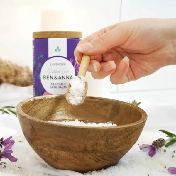 Ben & Anna bath salts in rose, packaged in a cardboard box with natural cork lid, shown alongside a wooden bowl with the bath salts inside and a spoonful being scooped out