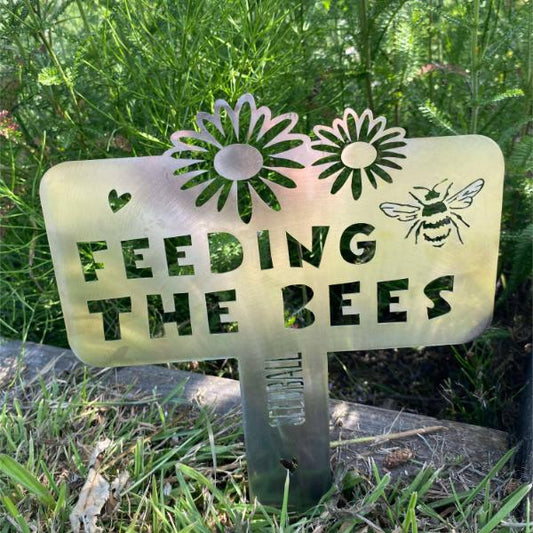 "Feeding the Bees" stainless steel sign by Seedball, with bee and floral cutout design shown planted in grass.