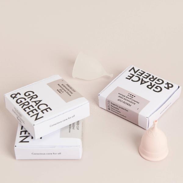 Two white boxes of Grace & Green menstrual cups arranged on a light beige background, with a translucent silicone cup and a pale pink menstrual cup placed beside them.