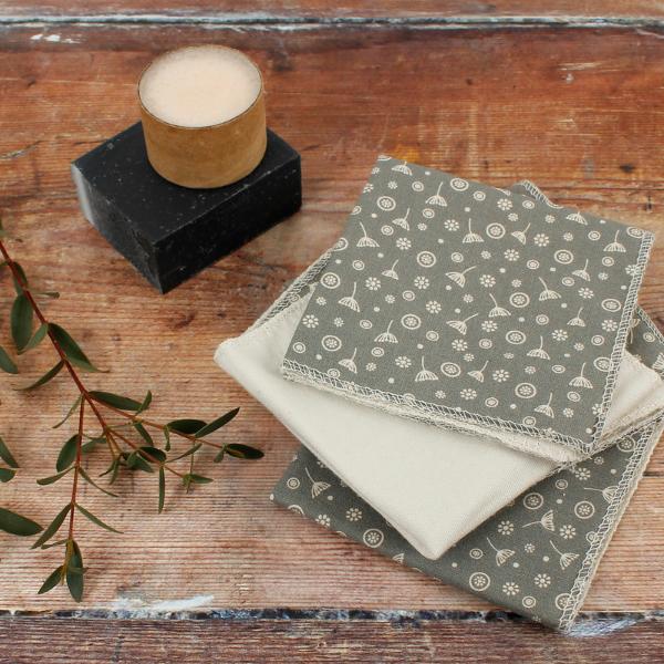 A stack of folded reusable wipes or cloths in muted grey and cream tones with a small repeating motif, arranged on a rustic wooden surface beside a candle and greenery.