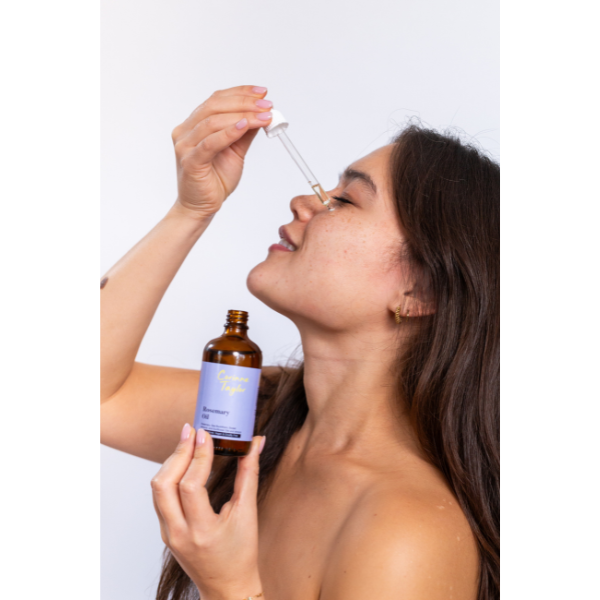 Rosemary oil for skin and hair in glass bottle being held in a model's hand with oil being applied to face with white pipette