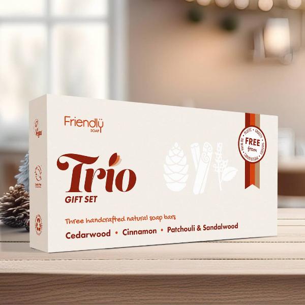 A white box of Friendly Soap Trio Gift Set, featuring natural soaps: Cedarwood, Cinnamon, and Patchouli & Sandalwood. The box is displayed on a wooden surface with a blurred warm indoor background.