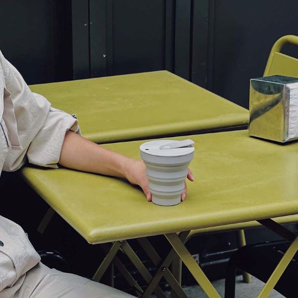 Collapsible cup shown in warm grey in a person's hand sitting at an outside bistro table 