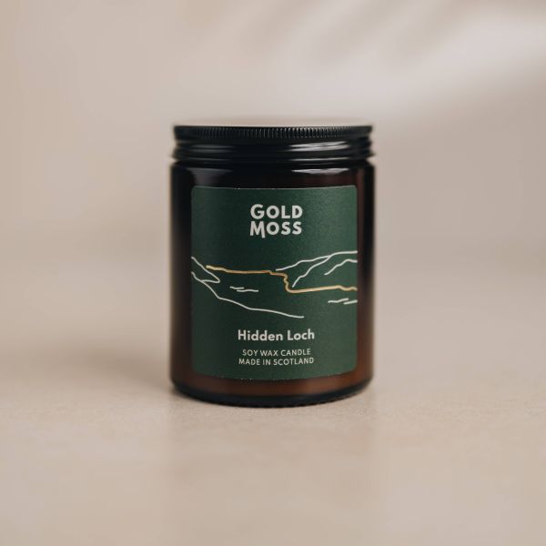 An amber glass jar candle with a black lid and a dark green label reading 'GOLD MOSS' and 'Hidden Loch', photographed on a warm beige surface with a softly lit neutral background.