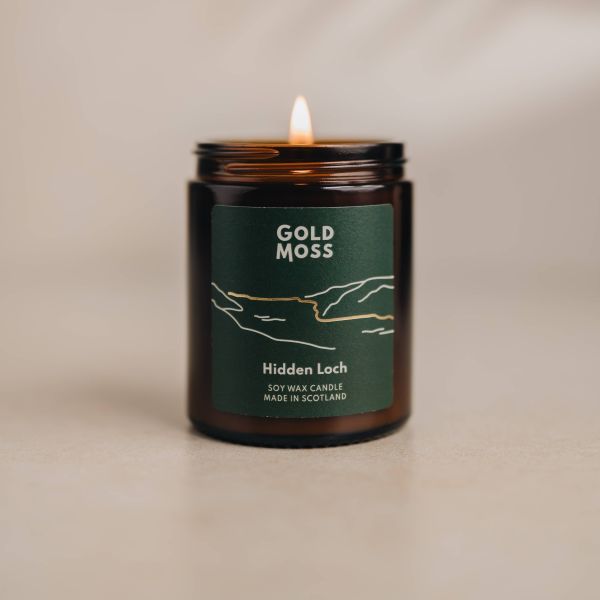 A lit amber glass jar candle with a dark green label reading 'GOLD MOSS' and 'Hidden Loch', shown on a warm beige surface against a softly lit neutral background.