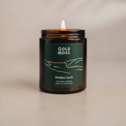 A lit amber glass jar candle with a dark green label reading 'GOLD MOSS' and 'Hidden Loch', shown on a warm beige surface against a softly lit neutral background.