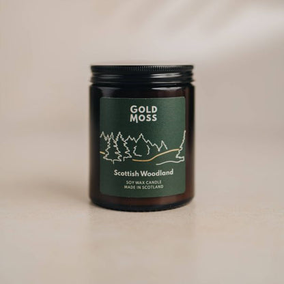 A dark amber glass jar candle with a black metal lid and a dark green label reading 'GOLD MOSS' and 'Scottish Woodland', photographed on a warm beige surface against a matching neutral background.