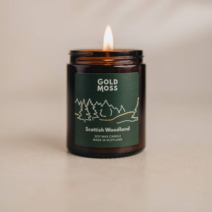 A lit amber glass jar candle labeled 'Gold Moss Scottish Woodland' on a neutral beige surface with a soft beige background.