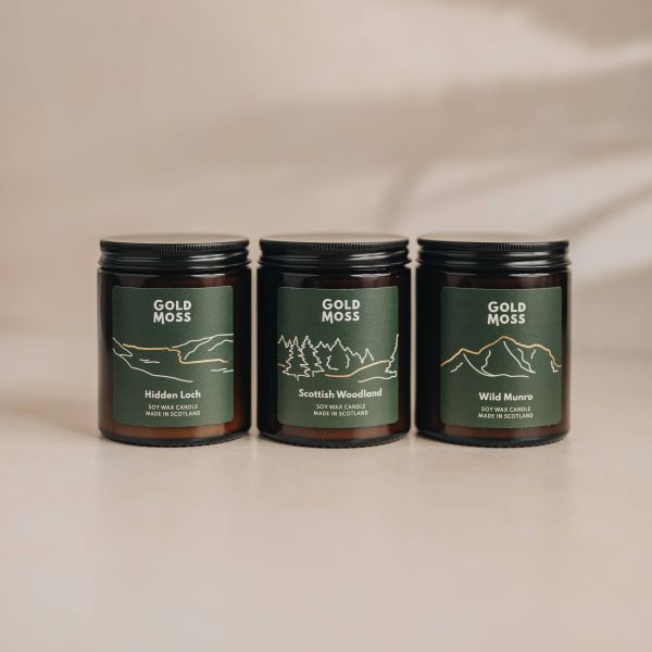 Three amber glass soy wax candles with black lids and dark green labels reading Gold Moss, arranged on a light beige surface against a soft beige background. The labels show simple line drawings and the scents Hidden Loch, Scottish Woodland, and Wild Munro.