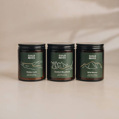 Three amber glass soy wax candles with black lids and dark green labels reading Gold Moss, arranged on a light beige surface against a soft beige background. The labels show simple line drawings and the scents Hidden Loch, Scottish Woodland, and Wild Munro.