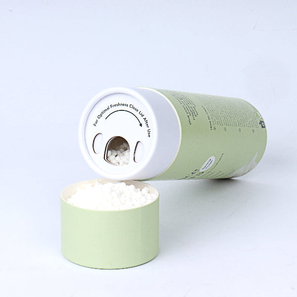 Green cardboard cylinder box containing eco fragrance booster, showing side on with beads inside. Top of box reads 'For optimal freshness close lid after use' with an arrow