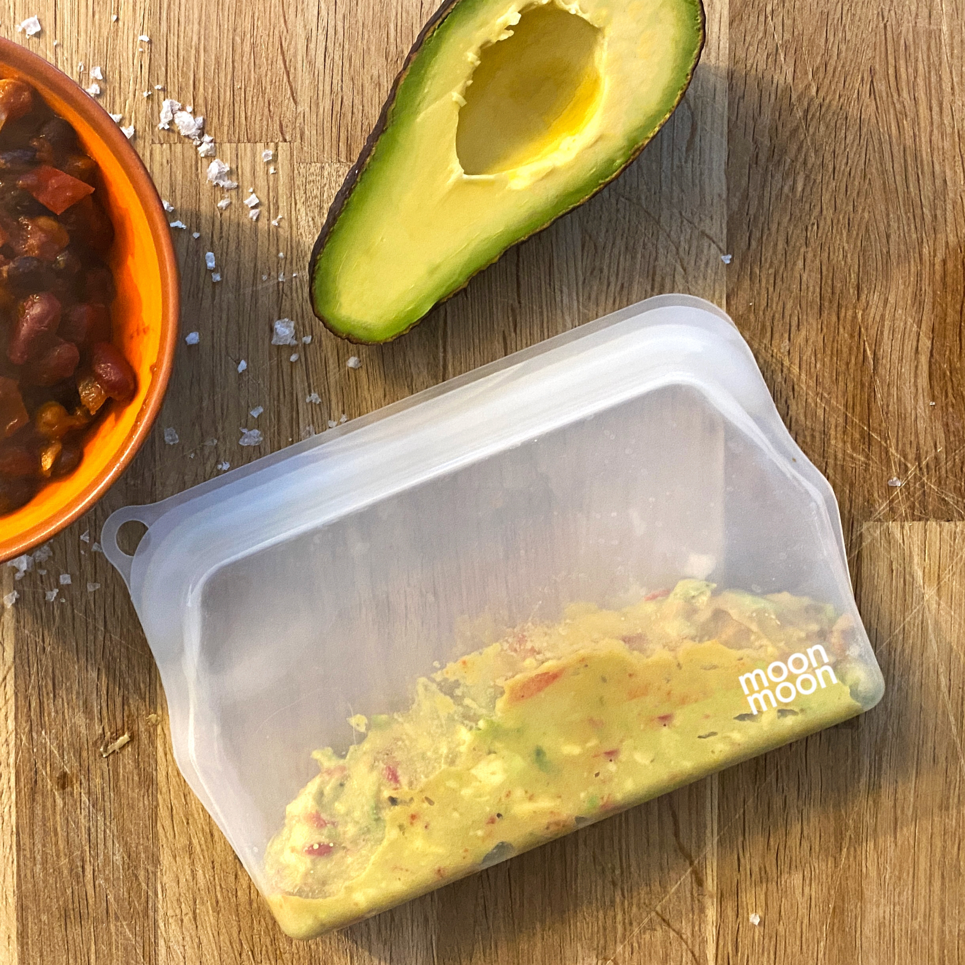 Silicone pouch of guacamole with an avocado half and salsa on a wooden surface, featuring the moonmoon brand.