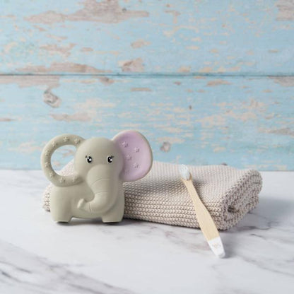 An elephant-shaped baby teether in a warm off-white colour, shown next to a knitted hand towel and baby toothbrush