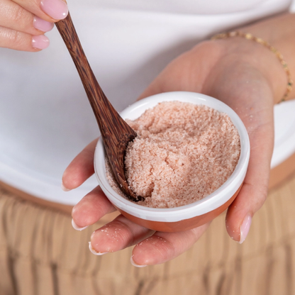 A close-up of a hand holding a small white bowl filled with pink bath salt crystals, with a wooden spoon lifting some of the salt. 