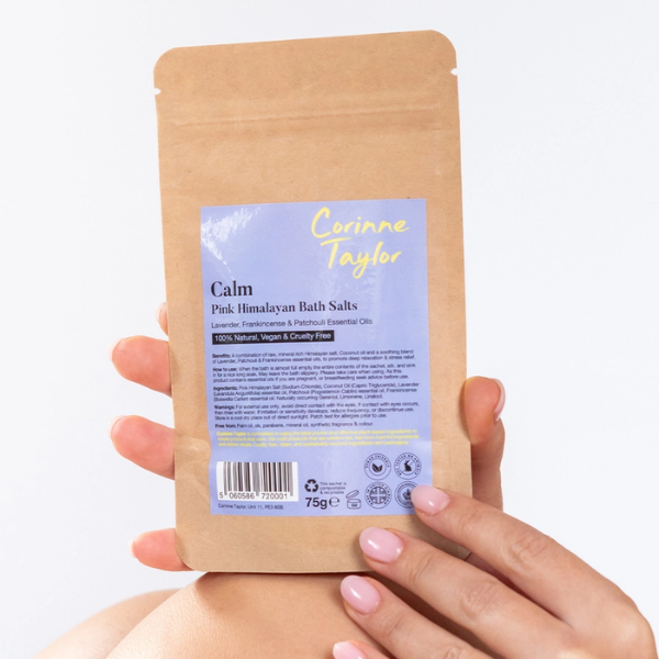 Hands holding a brown kraft paper pouch of Corinne Taylor Calm Pink Himalayan Bath Salts against a clean white background.