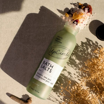 Glass bottle of UpCircle Bath Salts on a textured surface with dried flowers and leaves.