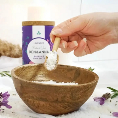 Ben & Anna bath salts in rose, packaged in a cardboard box with natural cork lid, shown alongside a wooden bowl with the bath salts inside and a spoonful being scooped out