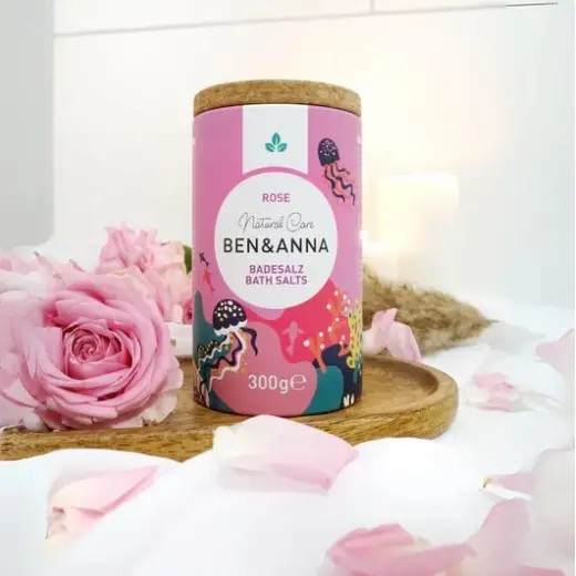 Ben & Anna bath salts in rose, packaged in a cardboard box with natural cork lid shown next to some pink roses