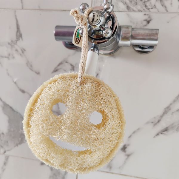 A round natural loofah sponge with a smiley face cutout hangs from a metal faucet handle in a bathroom setting with marble tiles.