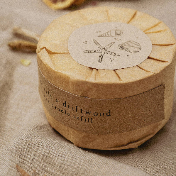 Round candle refill with natural design on a textured surface with seashells and driftwood and label reading "sea salt and driftwood soy wax candle 