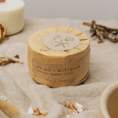 Round candle refill with natural design on a textured surface with seashells and driftwood and label reading "sea salt and driftwood soy wax candle refill"