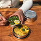 Stainless steel stackable round lunch boxes with food with a person dipping cutlery inside, on a wooden surface.