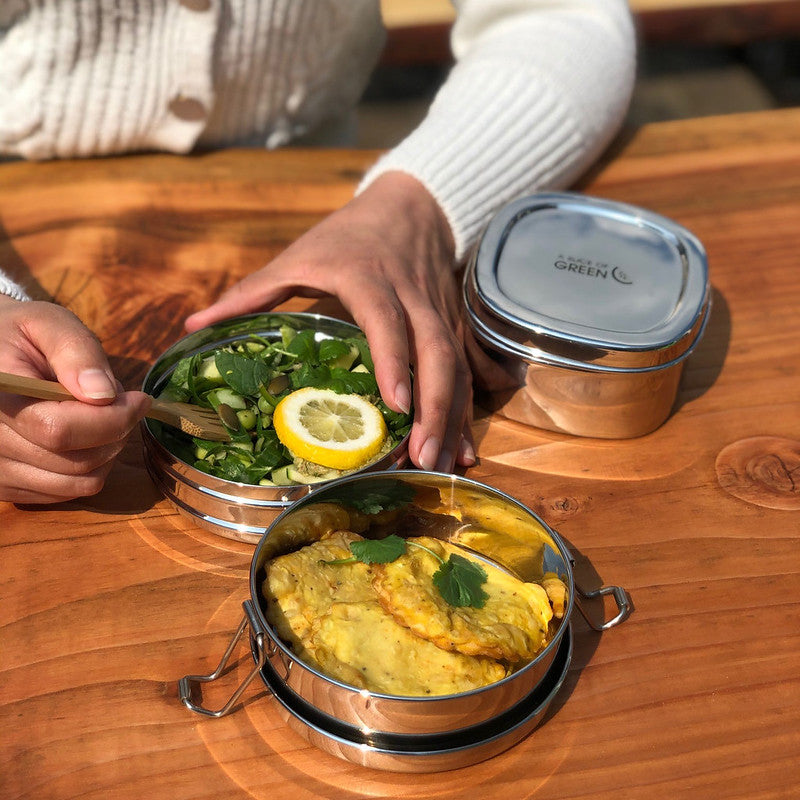 Stainless steel stackable round lunch boxes with food with a person dipping cutlery inside, on a wooden surface.