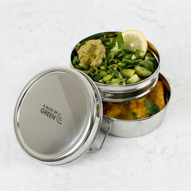 Stainless steel stackable round lunch boxes shown open with food inside on a white surface, featuring the brand 'A Slice of Green'.