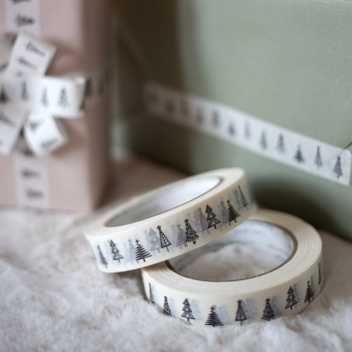 Two rolls of white paper tape with a repeating black Christmas tree pattern on a white background, styled on a white faux fur surface with gift boxes in the background.