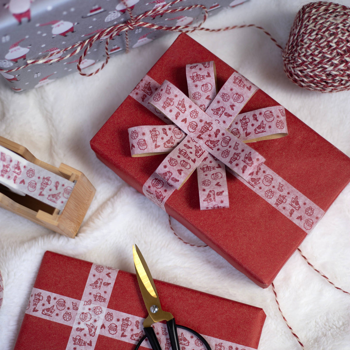 Red wrapping paper with white paper tape with red Santas shaped into a bow, styled on a white textured surface with scissors, a spool of twine.