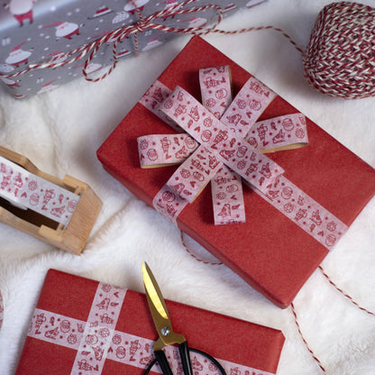 Red wrapping paper with white paper tape with red Santas shaped into a bow, styled on a white textured surface with scissors, a spool of twine.