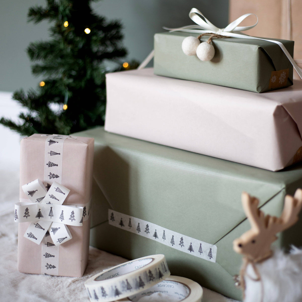 Three wrapped Christmas gift boxes in sage green and blush pink paper, styled with patterned ribbons and paper tape, with a blurred Christmas tree in the background.