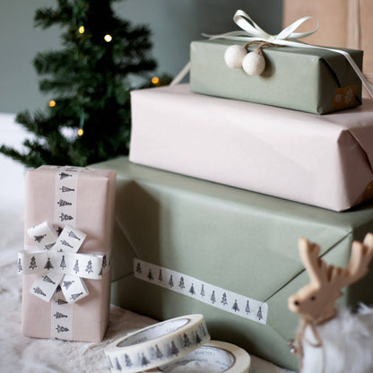 Three wrapped Christmas gift boxes in sage green and blush pink paper, styled with patterned ribbons and paper tape, with a blurred Christmas tree in the background.