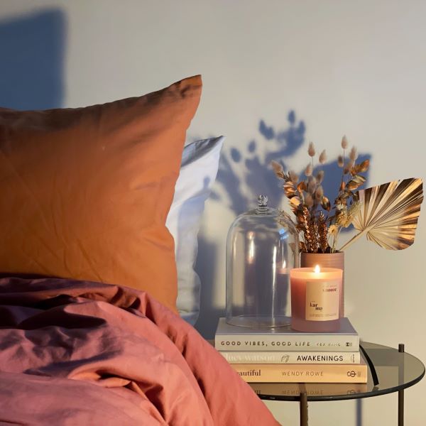 Clean-burning refillable mini Christmas candle in Post Roast Snooze fragrance, in an orange glass refillable jar, shown lit next to a bed