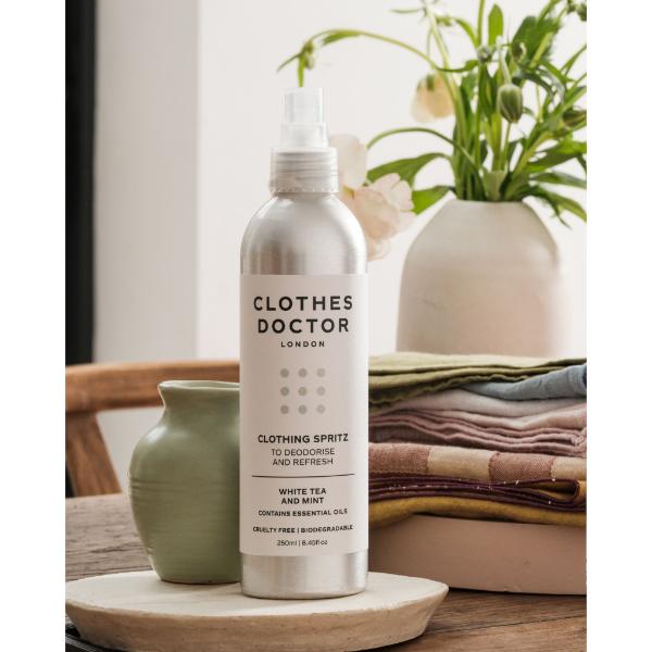 Clothes Doctor Clothing Spritz in an aluminium bottle with spray top on a worktop next to some flowers and cloth napkins