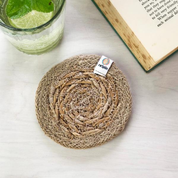 Round seagrass woven coaster with jute trim, on a marble surface with a ReSpiin brand label, next to a book and glass.