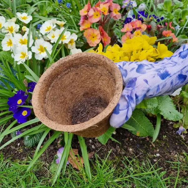 Coco coir biodegradable and reusable plant pot shown in a hand next to some flowering plants