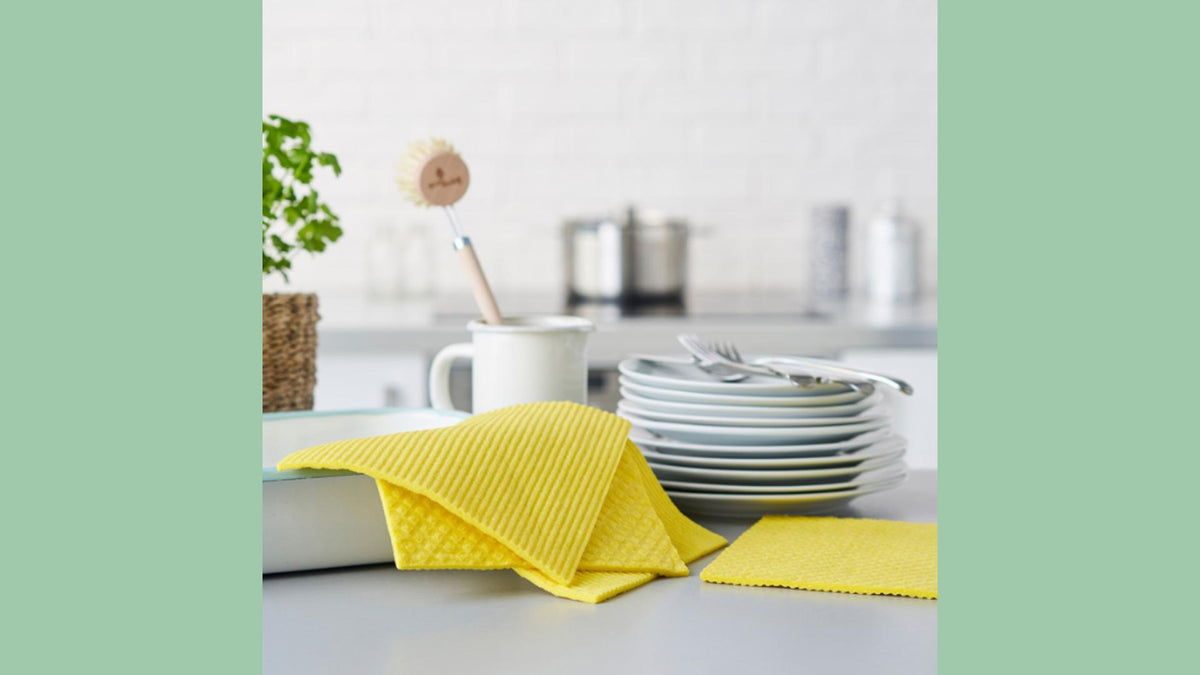 A set of yellow compostable dishcloths arranged on a kitchen counter next to a stack of white plates and a white mug with a plastic-free dishbrush.