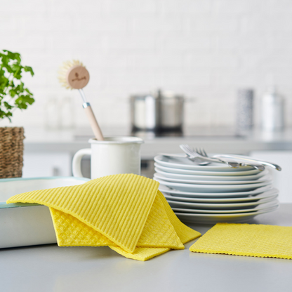 Compostable yellow sponge dishcloths on a kitchen counter with plates and utensils in the background