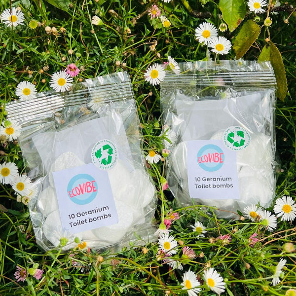Two packages of EcoVibe Geranium Toilet Bombs on grass with flowers