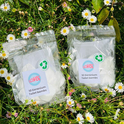 Two packages of EcoVibe Geranium Toilet Bombs on grass with flowers
