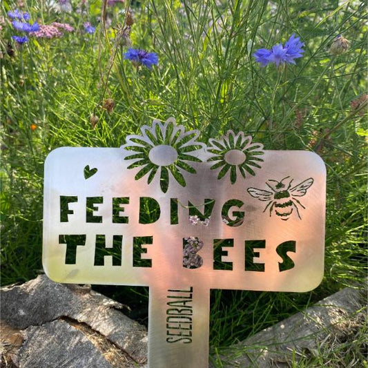 "Feeding the Bees" stainless steel sign by Seedball, with bee and floral cutout design shown next to wildflowers.