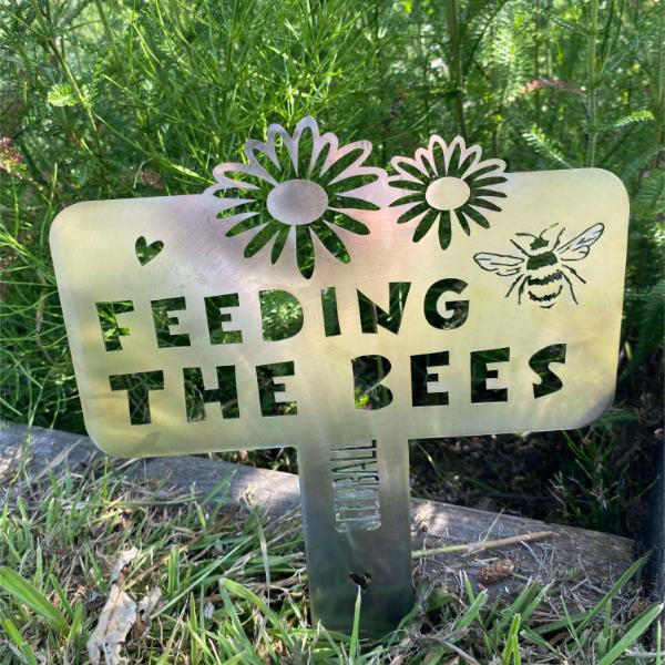 "Feeding the Bees" stainless steel sign by Seedball, with bee and floral cutout design shown planted in grass.