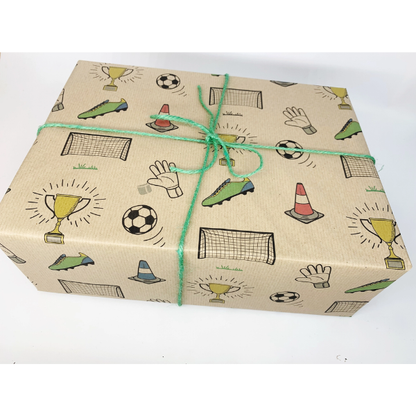 A rectangular gift box wrapped in brown kraft wrapping paper printed with football-themed illustrations including trophies, soccer balls, goals and a goalkeeper hand, tied with a green string on a white background.