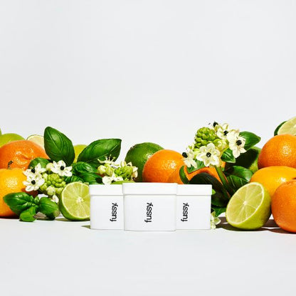 Three Fussy deodorant refills surrounded by citrus fruits and flowers on a white background