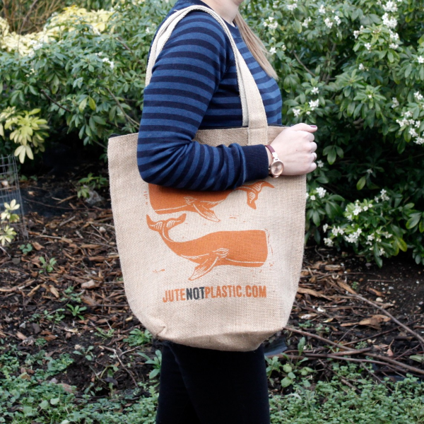 Person holding a jute tote bag with an orange whale design and 'Jute Not Plastic' text in a garden setting.