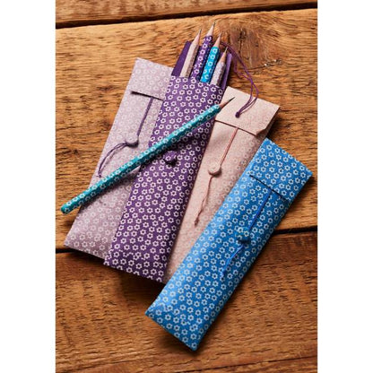 Three paper pencil cases in light purple, pink, and blue with a white floral pattern, shown on a rustic wooden table with pencils inside.