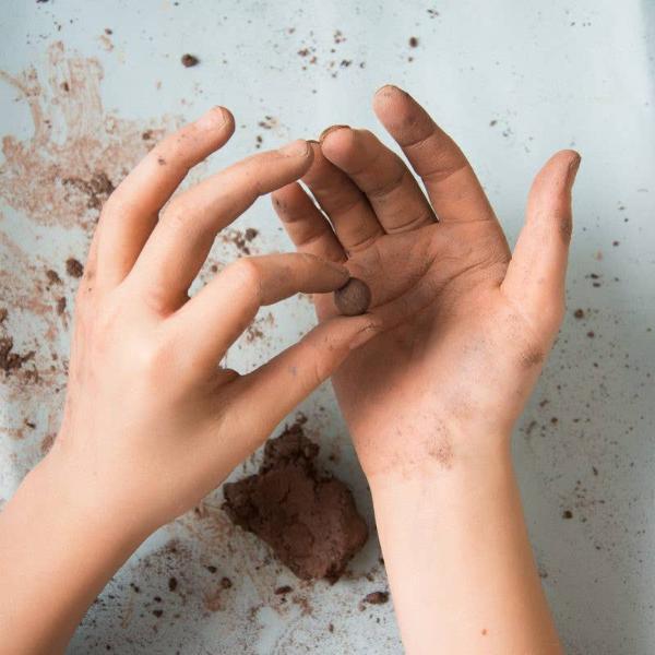 Hands holding a small ball of brown clay making a seedball on a textured surface