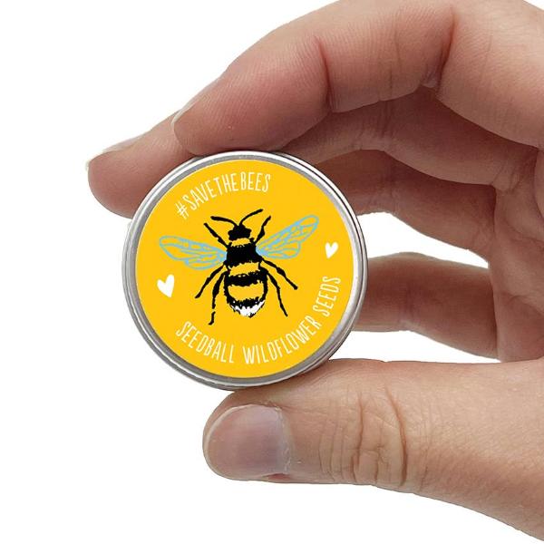 Hand holding a round yellow seedball tin with a bee design and text reading "#Savethebees Seedball wildflower seeds" on a white background